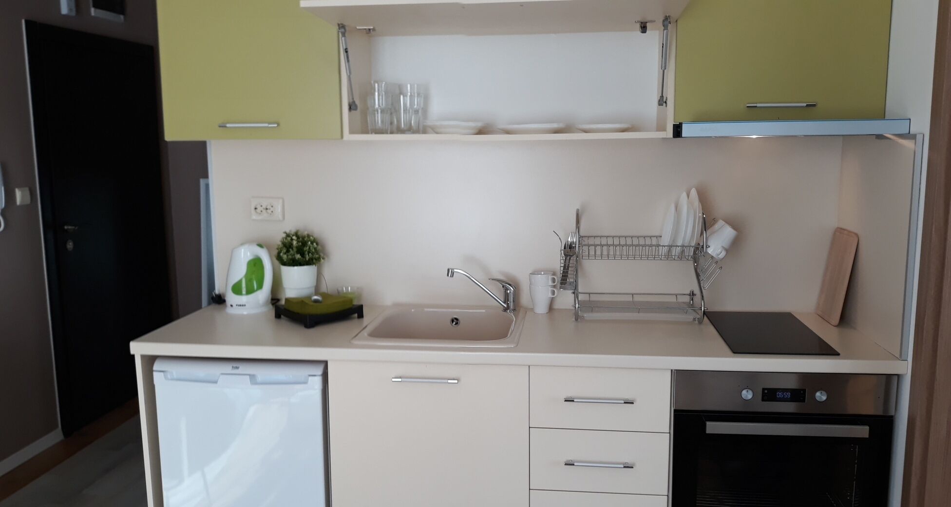 Eine kompakte Küche mit Geschirrspüler, Spüle und kleinem Backofen. Über der Spüle befinden sich Schränke und eine Arbeitsplatte mit einem Geschirrgestell.