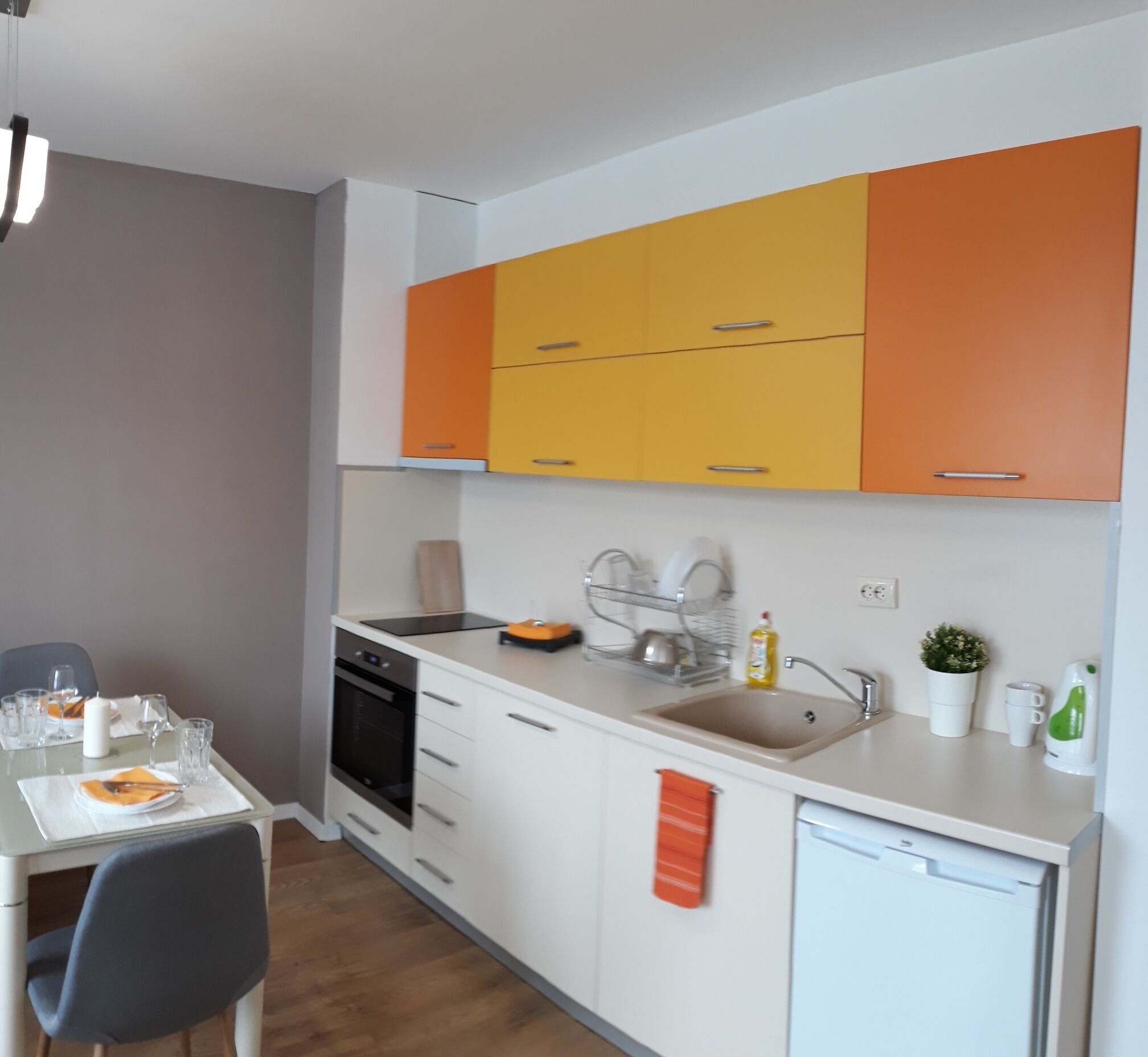 Una cocina compacta con armarios blancos y naranjas, un lavavajillas, un fregadero y una zona de comedor con una mesa preparada para dos.