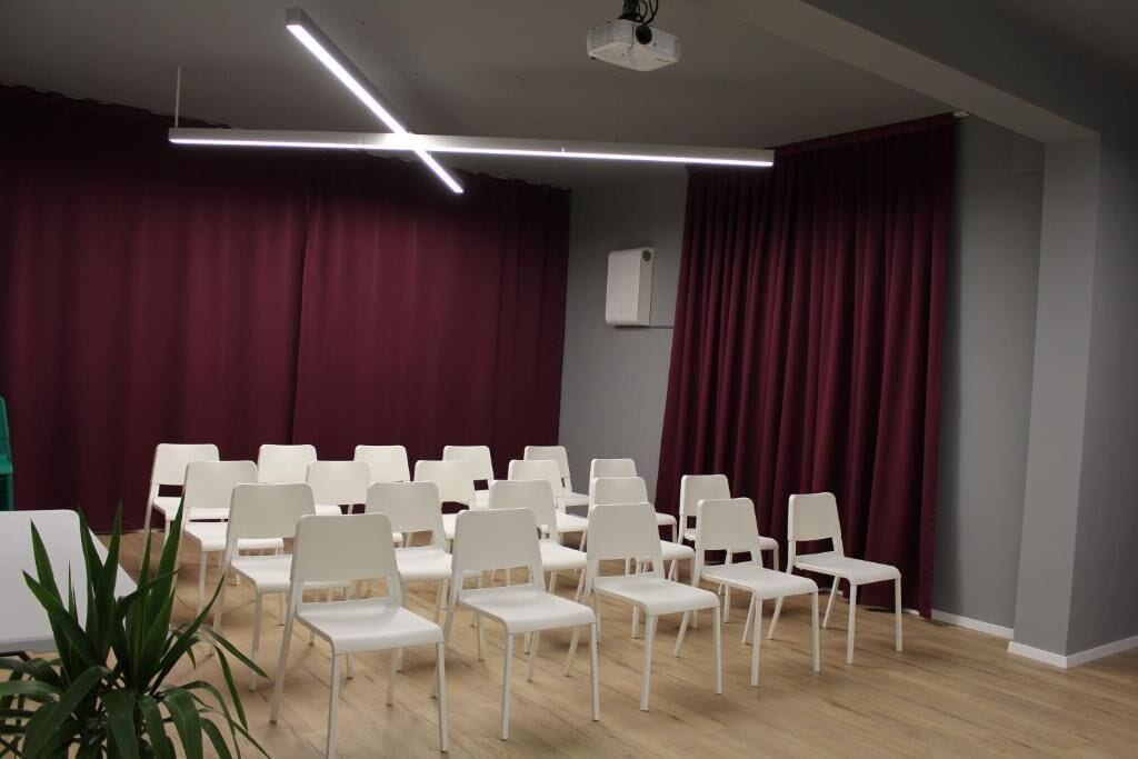 Un salón de conferencias con sillas blancas dispuestas en filas, un proyector en el techo y un telón rojo de fondo.