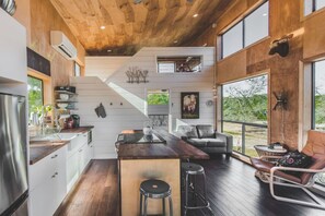 Open-plan living space with a kitchen and seating area, featuring a staircase and large window with outdoor views.