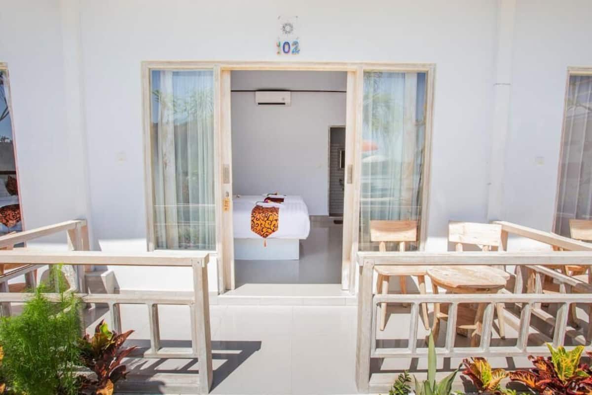 A hotel room with a bed, a small table, and a chair. There are two doors leading to the room, and a balcony with a railing and potted plants.
