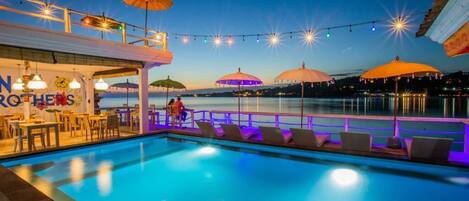 A poolside area with umbrellas, string lights, and a bar/lounge area with a sign reading "N Brothers."