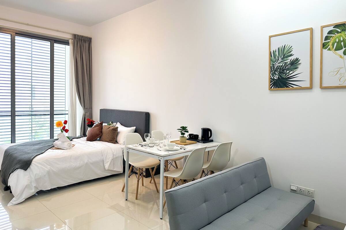 A modern hotel room with a bed, a dining table with chairs, a sofa, and a small table with a kettle and cups.