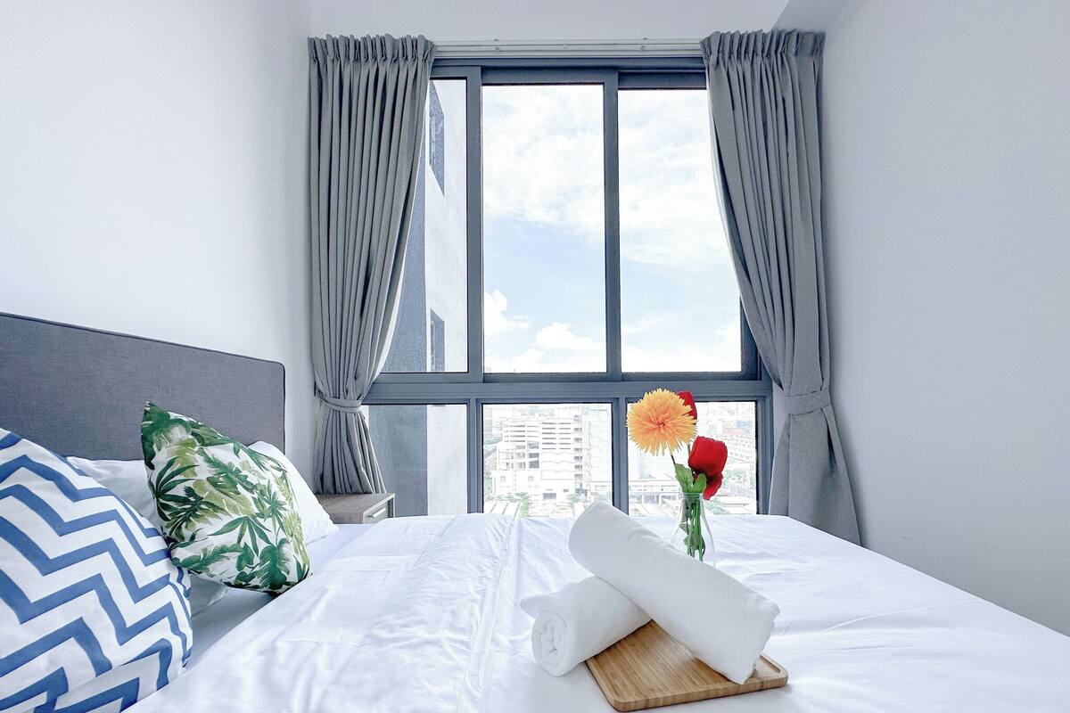 A hotel room with a large bed, a view of the city through a window, and a vase with flowers on the nightstand.