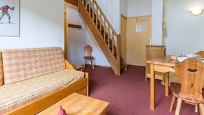 A cozy room with a wooden staircase, a sofa, a dining table with chairs, and a small wooden cabinet. - Duplex 3-room, 8 pers, balcony, near slopes (Le Corbier)