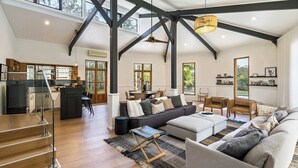A spacious living area with a high ceiling, wooden staircase, and a kitchen in the background. - The Noosa Ranch - Where Nature meets Luxury (Tinbeerwah)