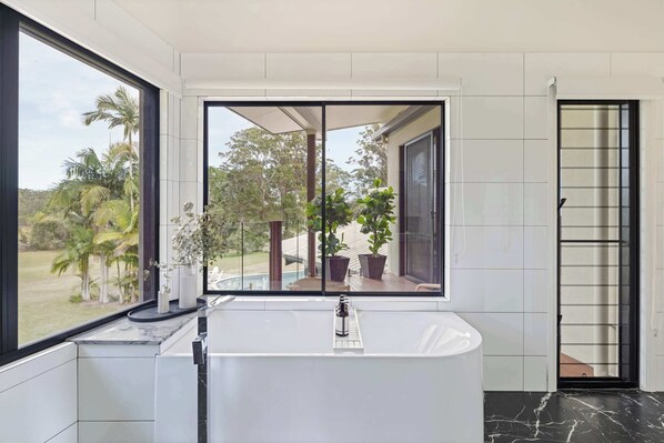 Un baño moderno con una bañera grande, una ventana con vista a follaje y una puerta con celosía de metal.