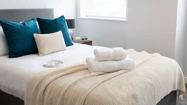 A neatly made bed with a white and beige blanket, teal and white pillows, and a bedside table with a lamp and books. - Contemporary 1 Bedroom Liverpool Apartment (Liverpool)
