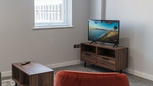 Ein Wohnzimmer mit Fernseher auf einem Holzständer, einem kleinen Tisch mit Fernbedienung und einem Fenster mit Jalousien.