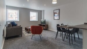 Ein modernes Wohnzimmer mit einem grauen Sofa, einem roten Sessel, einem Esstisch mit Stühlen, einem Fernseher und einem gerahmten Bild an der Wand.