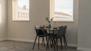 Un coin repas avec une table dressée pour quatre personnes, deux fenêtres équipées de stores et une vue sur les bâtiments environnants.