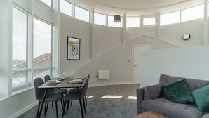 Uma sala de estar moderna com área de jantar, grandes janelas, uma escada e um sofá cinza com almofadas verdes.