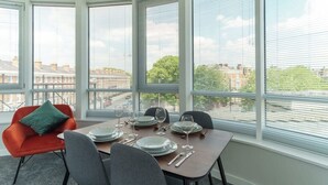 Uma sala de jantar com mesa posta para quatro pessoas, janelas amplas e vista para a paisagem urbana.