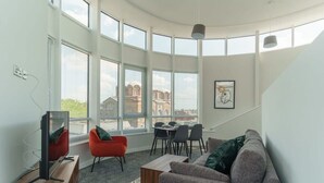 Uma sala de estar moderna com uma parede curva, janelas amplas, uma televisão, um sofá, uma mesa de jantar com cadeiras e uma escada.