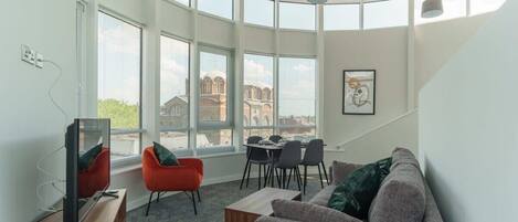 Una sala moderna con una pared curva, ventanas amplias, un televisor, un sofá, una mesa de comedor con sillas y una escalera.