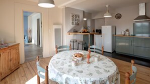 Ein gemütlicher Essbereich mit einem runden Tisch, Stühlen und einer Tischdecke mit Blumenmuster. Im Hintergrund ist eine Küche mit Kühlschrank, Mikrowelle und Herd zu sehen.