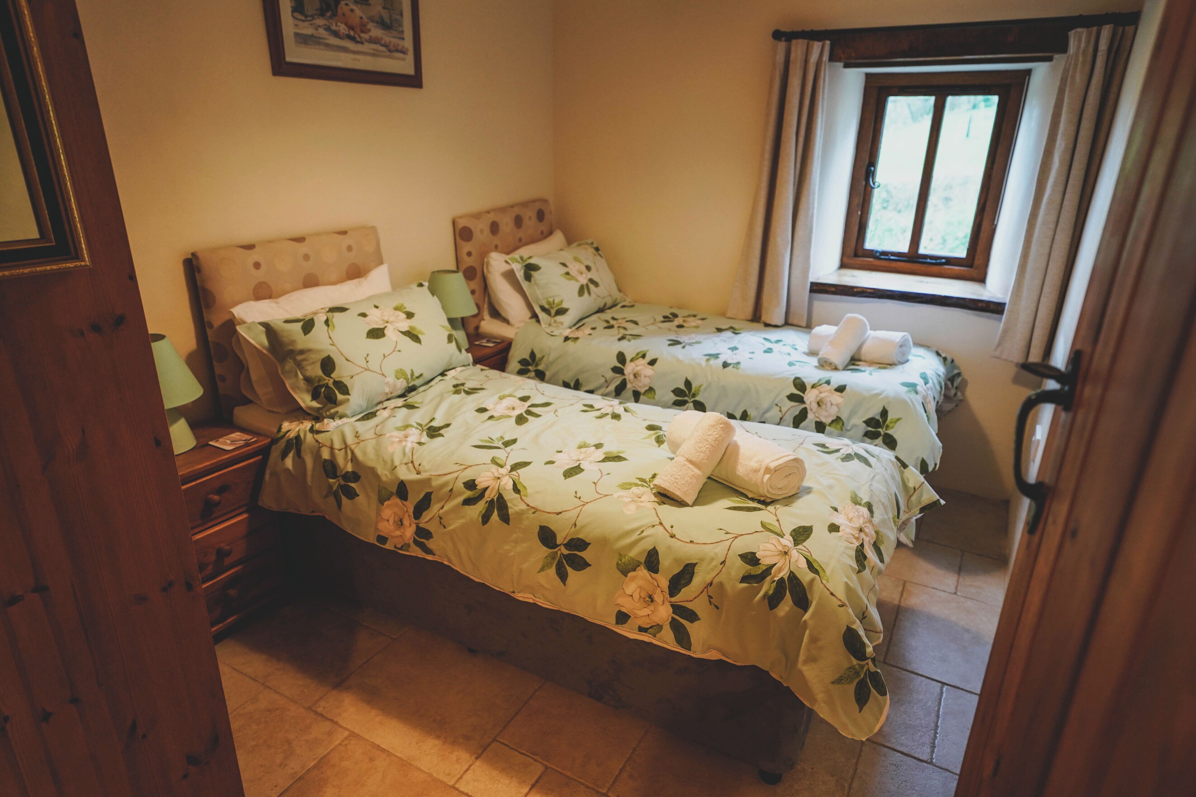 Une chambre avec deux lits simples, chacun agrémenté de draps à motifs floraux et d’une serviette roulée posée sur le lit. Il y a une table de chevet avec une lampe et une porte en bois sur le côté gauche.