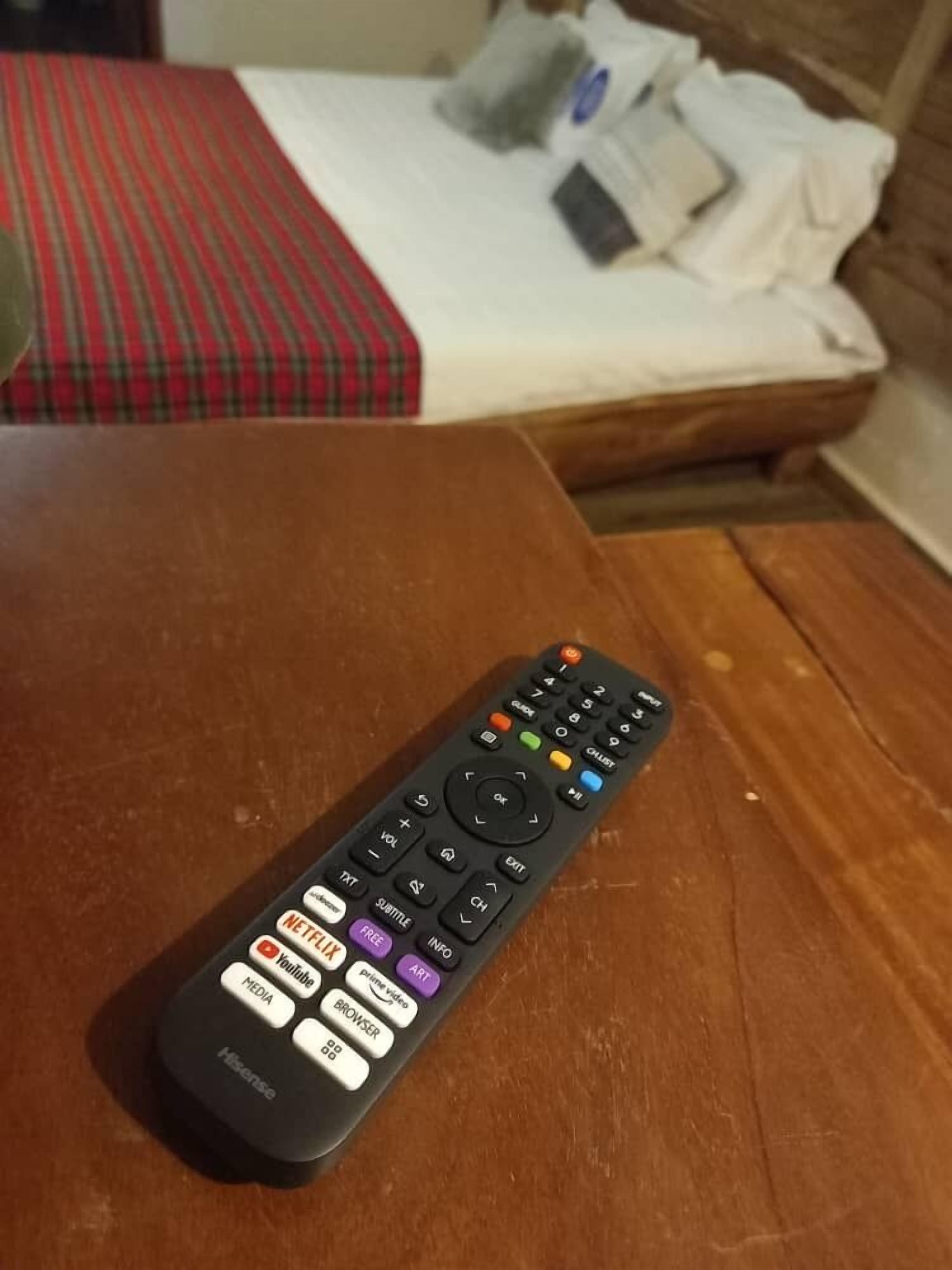 A remote control on a wooden table with a bed in the background.
