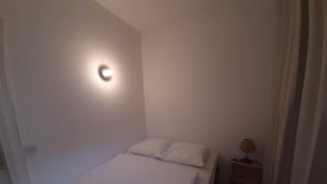Ein kleines Zimmer mit einem Einzelbett, einem Nachttisch, einer Wandleuchte und einem Vorhang.