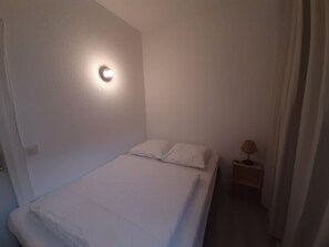 A small, single-bed room with a bedside table, a wall-mounted light, and a curtain.