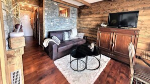 A cozy living room with a stone wall, wooden beams, a leather sofa, a TV on a wooden cabinet, and a white rug.