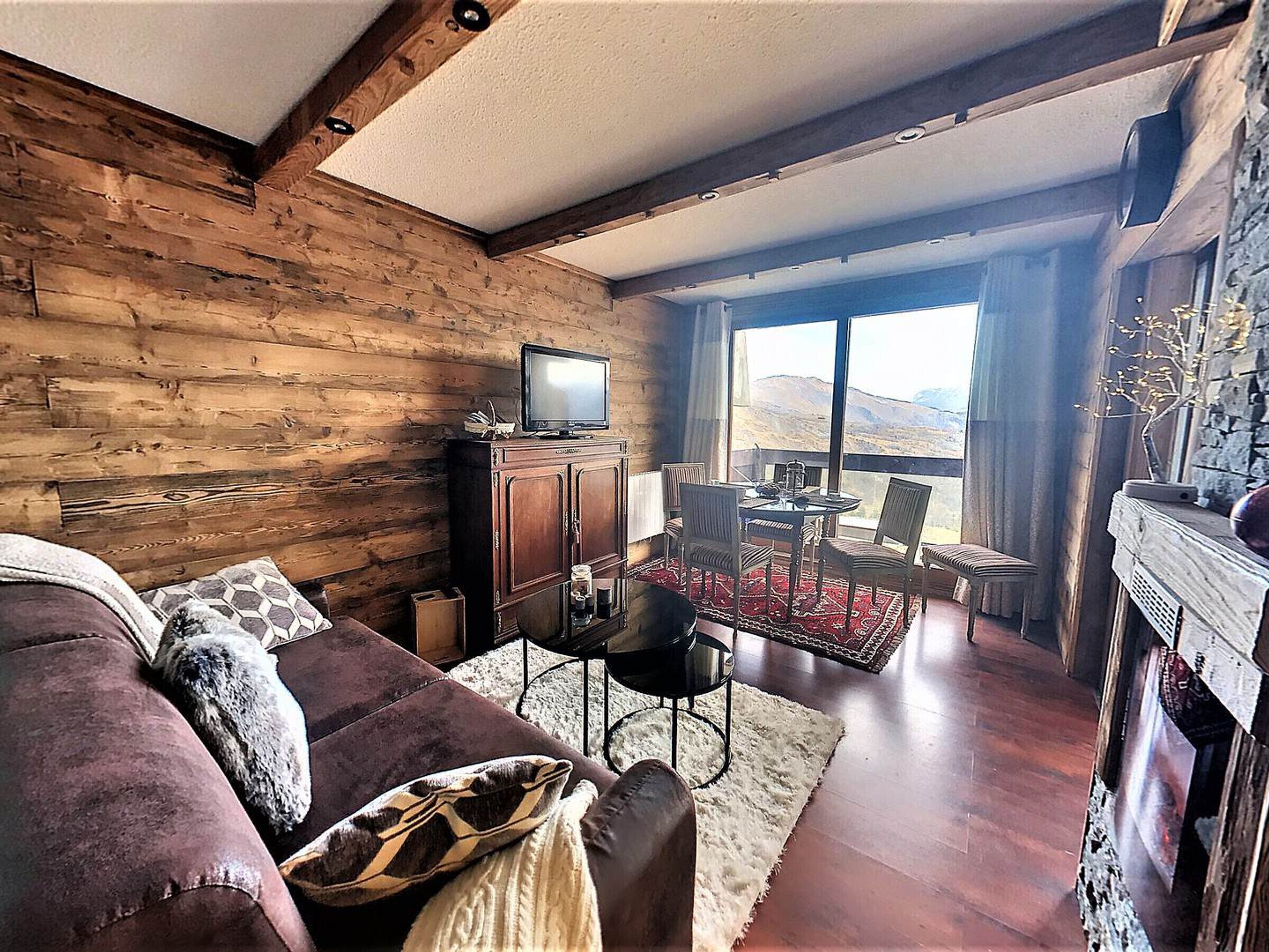 Un salon chaleureux avec un décor intérieur en bois, un téléviseur posé sur un meuble, un canapé avec des coussins, un coin repas avec des chaises et une table, et une vue sur les montagnes à travers de grandes fenêtres.