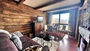 A cozy living room with a wooden interior, a television on a cabinet, a sofa with cushions, a dining area with chairs and a table, and a view of the mountains through large windows.