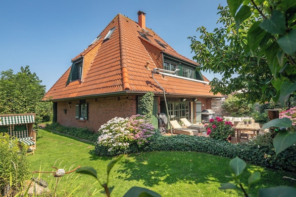 Une maison à deux étages, dotée d’un toit en pente, d’une façade en briques et d’un jardin bien entretenu avec une pelouse, des fleurs et du mobilier d’extérieur.