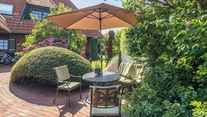 An outdoor patio with a round table and two chairs, surrounded by lush greenery and a large umbrella. - // Liebeney - Möwennest // (Norderney)