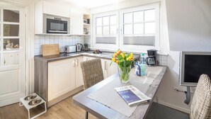 A modern kitchen with white cabinets, a microwave, a kettle, and a coffee maker. There is a dining table with a magazine and a vase of flowers. - // Liebeney - Möwennest // (Norderney)