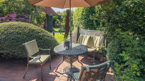 An outdoor patio with a round table and two chairs, surrounded by lush greenery and a large umbrella providing shade. - LIEBENEY - BEACH CHAIR // (Norderney)