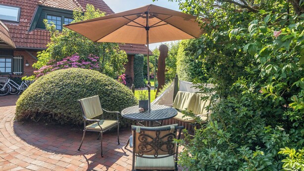 An outdoor patio with a round table and two chairs, surrounded by lush greenery and a large umbrella. - LIEBENEY - BEACH CHAIR // (Norderney)