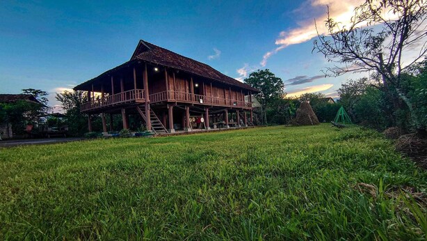 A wooden house with a balcony and a staircase leading to the upper floor, surrounded by a grassy field. - MUONG PHANG HOMESTAY (Muong Phang)