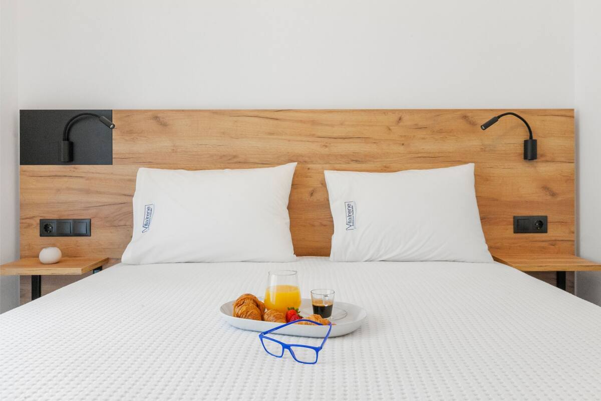 Un llit ben arropat amb matalàs blanc, dos coixins blancs, un capçal de fusta, làmpades laterals, una taula de nit amb una làmpada i un safata amb croissants, suc i vasos.