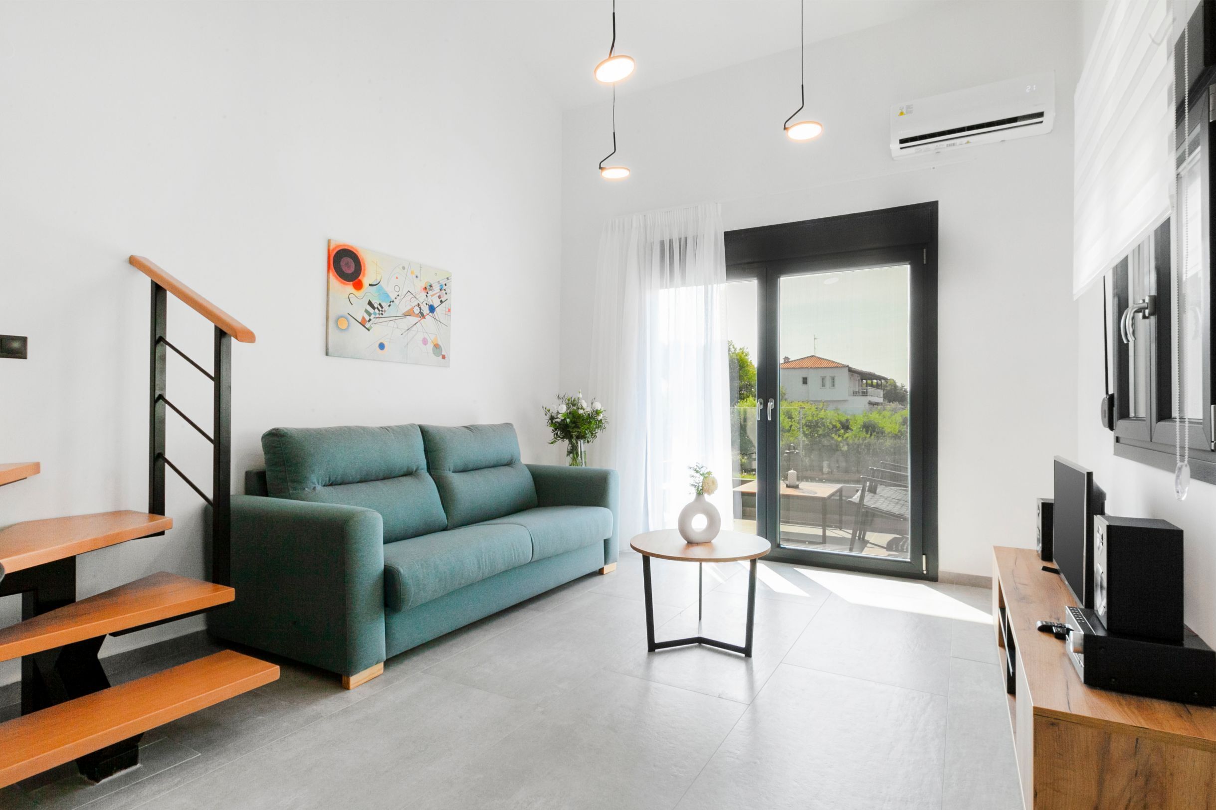 Una sala moderna con un sofá color turquesa, bancos de madera, una mesita redonda y un ventanal con vistas a una casa.