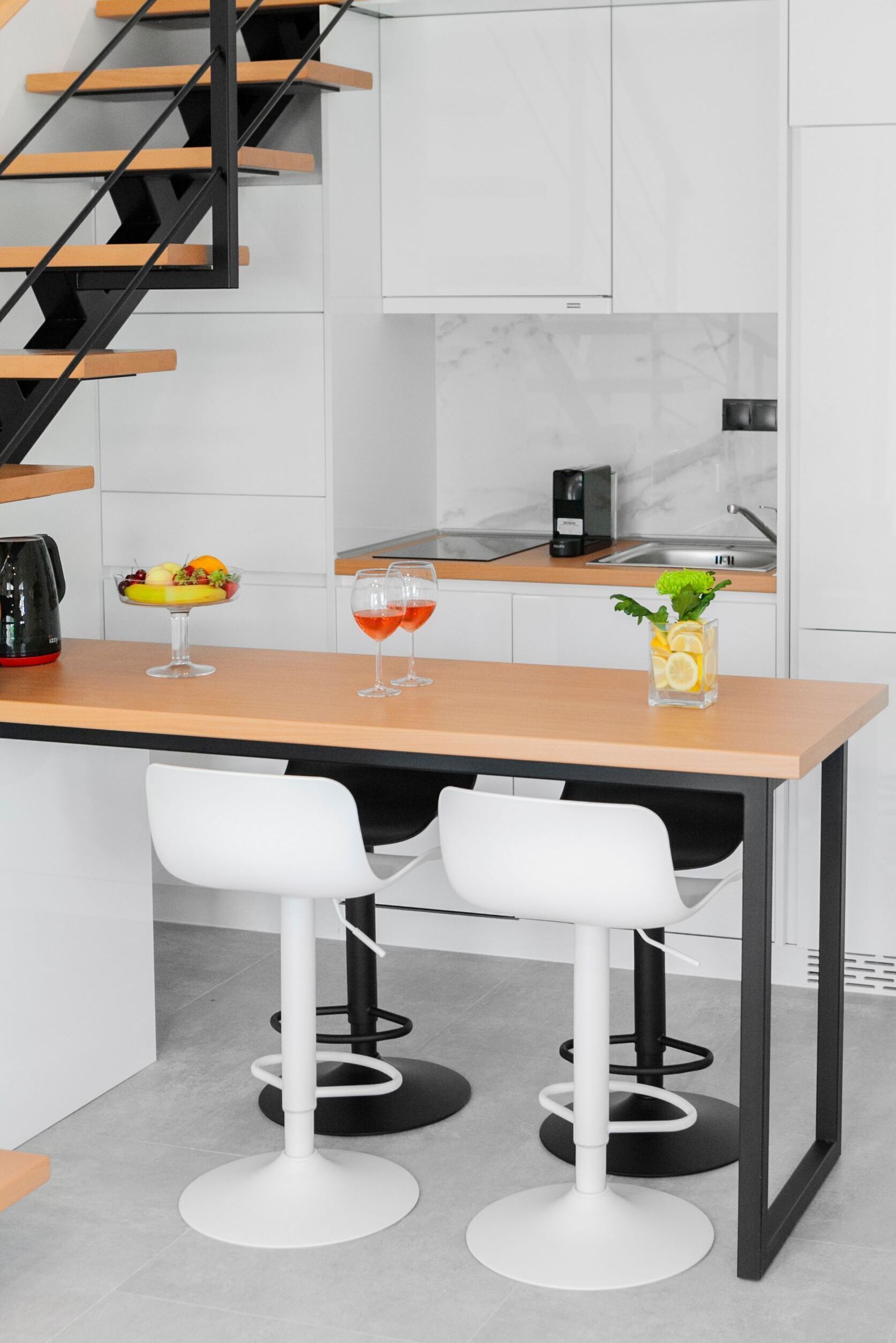 A modern kitchen with a wooden countertop, two white bar stools, a coffee maker, and a fruit bowl.