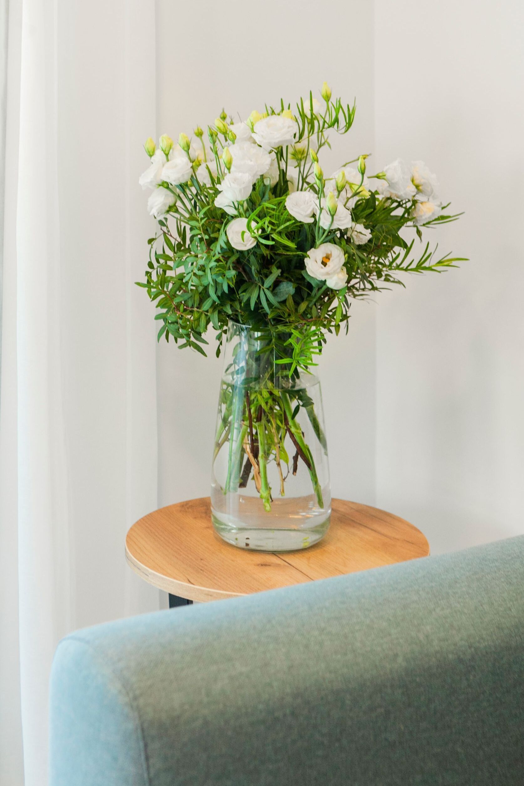 Un vase en verre transparent, orné de fleurs blanches et de feuilles vertes, est posé sur une table ronde en bois.