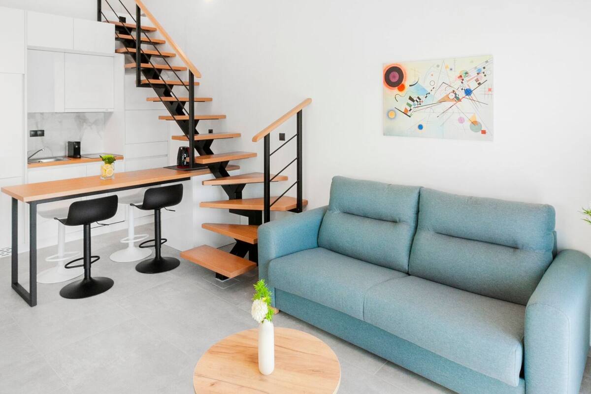 Un saló modern amb un sofà blau, una taula de centre rodona de fusta i una escala que porta a una zona de cuina.