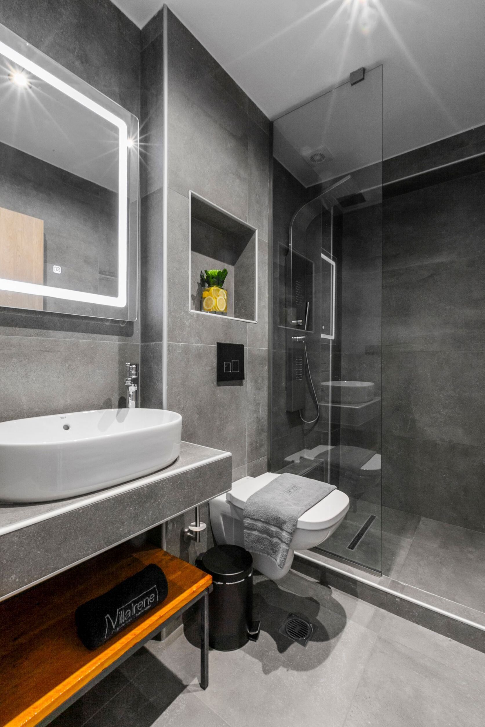 A modern bathroom with a large mirror, a white sink, a glass-enclosed shower, and a small shelf with a plant.