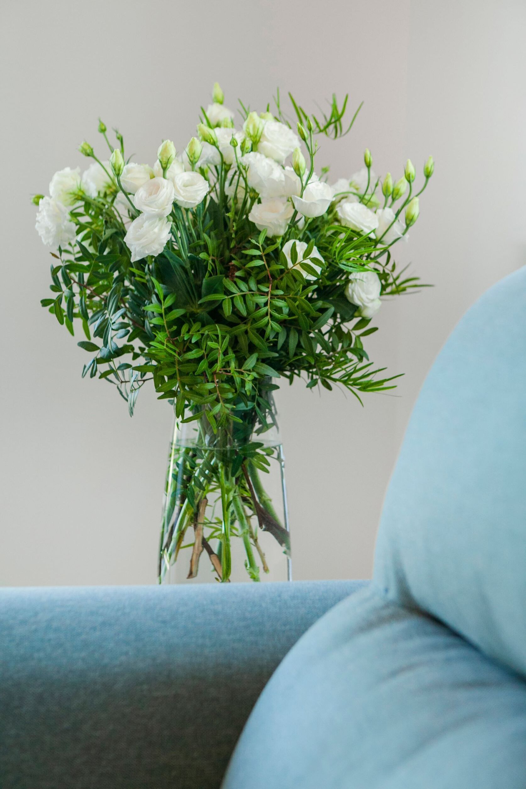 Eine transparente Glasvase mit weißen Blumen und grünen Blättern auf einer blauen Oberfläche.