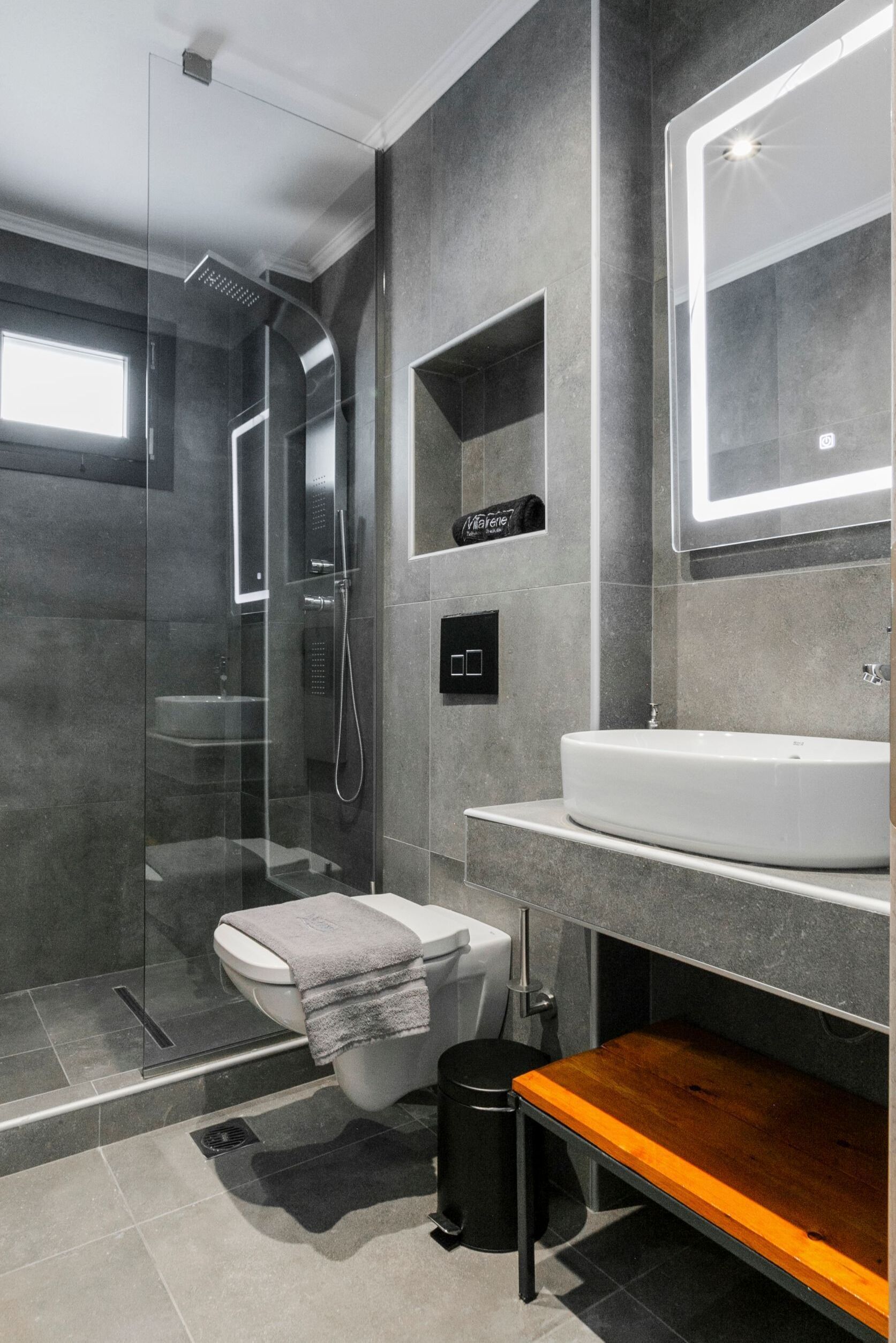 A modern bathroom with a glass-enclosed shower, a white sink, and a wooden bench.