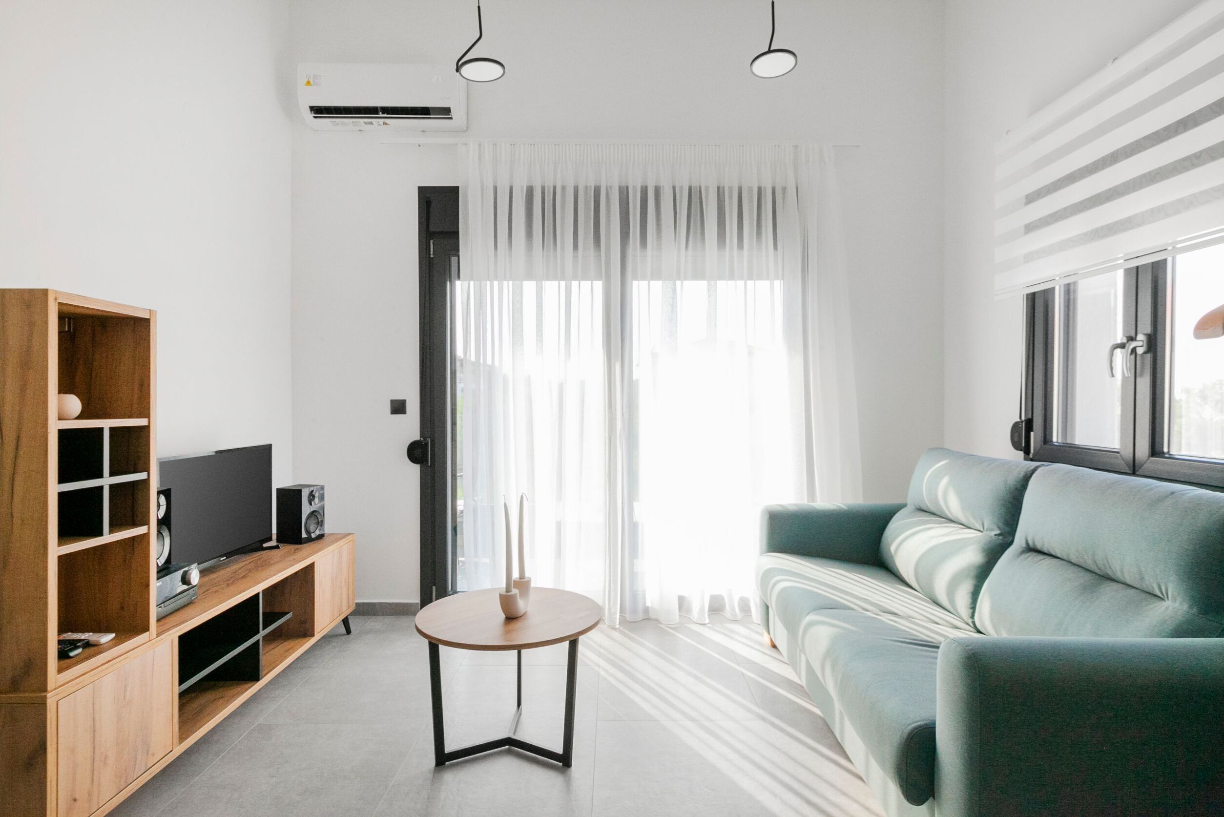 A modern living room with a wooden entertainment unit, a round table, a teal sofa, and a large window with sheer curtains.