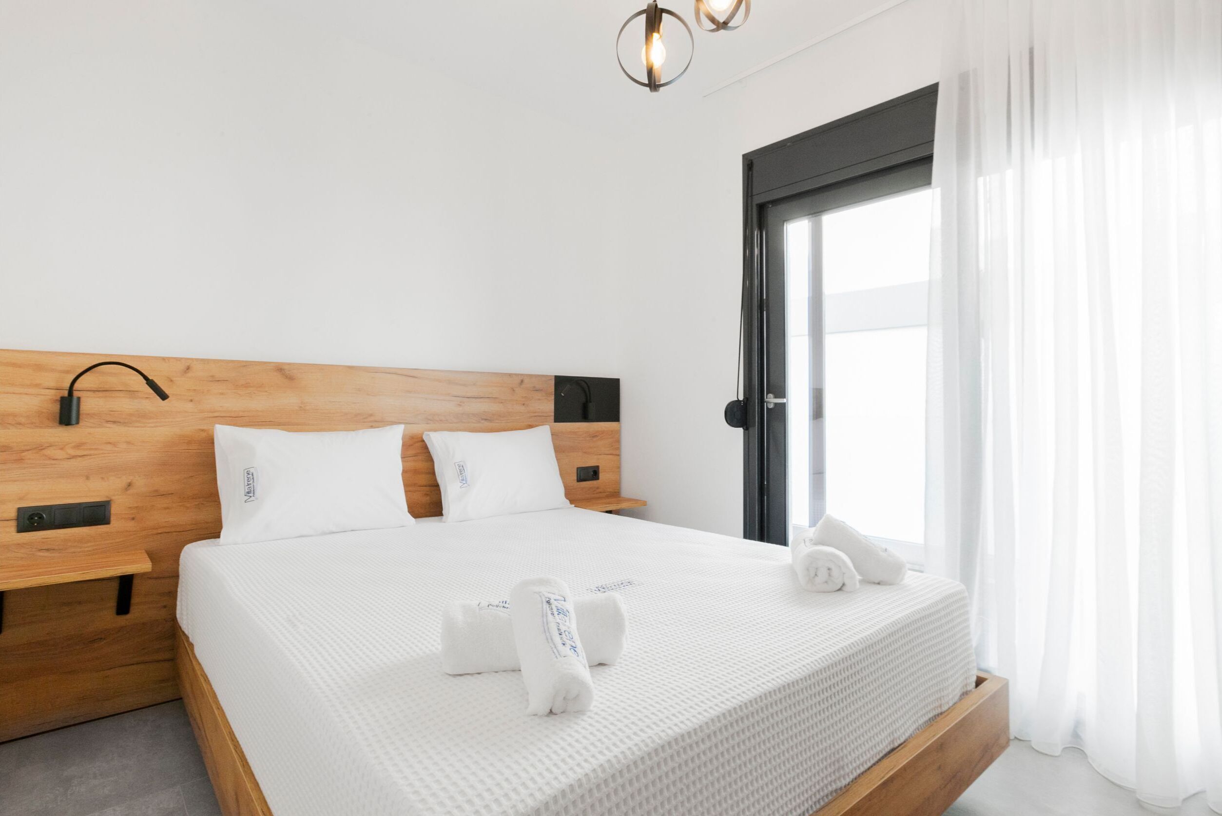 A neatly made bed with white bedding and two pillows, a wooden headboard, and a bedside table with a lamp.