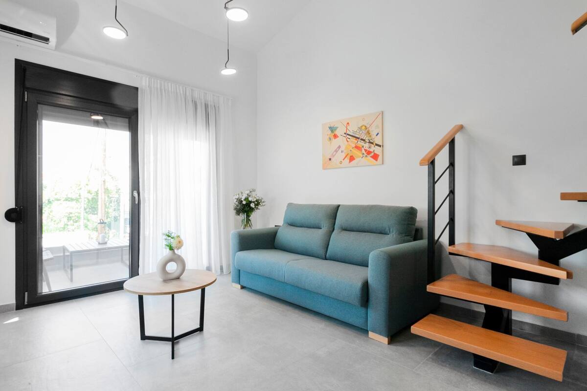 Un saló modern amb un sofà blau, una taula rodona petita amb un ram, una escala amb graons de fusta i un quadre a la paret.