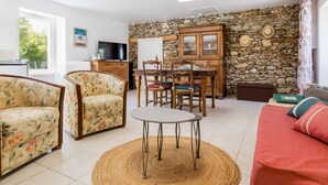 A living room with a stone wall, a television, a dining table with chairs, and a sofa. - Renovated Cottage Near Marshes, 12 km from Beach, Private Terrace, Garden, WiFi, Baby-Friendly (Villeneuve-en-Retz)