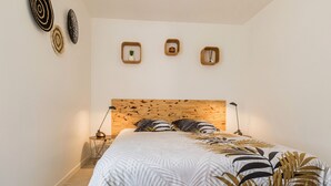 A bedroom with a wooden headboard, a bed with a leaf-patterned bedspread, two bedside tables with lamps, and three wall-mounted shelves with decorative items. - Renovated Cottage Near Marshes, 12 km from Beach, Private Terrace, Garden, WiFi, Baby-Friendly (Villeneuve-en-Retz)