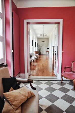 Corredor com piso xadrez, parede rosa e porta que dá para uma sala de jantar com soalho de madeira e cadeiras.