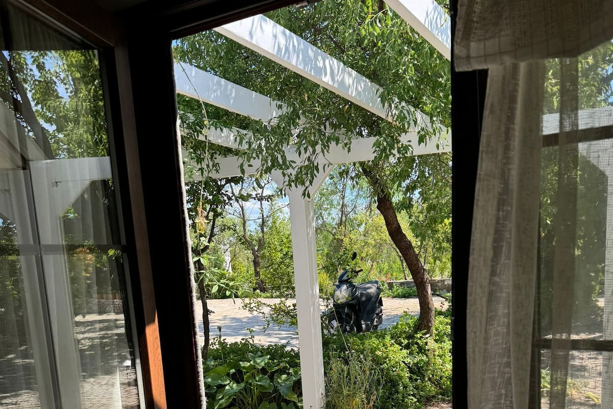 A view through a glass door into a garden area with a stone pathway, a white pergola, and a parked scooter.