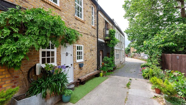 A narrow brick alley with a small garden area, a white door, and a wooden bench. - Harmonious Hideaway, by Veeve (London)