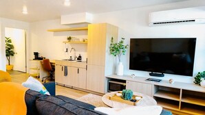 A modern living room with a sofa, coffee table, TV, and kitchen area with cabinets and a sink.
